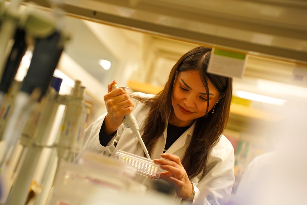 A researcher in a white lab coat uses a micropipette to transfer samples into a multiwell plate inside a laboratory.