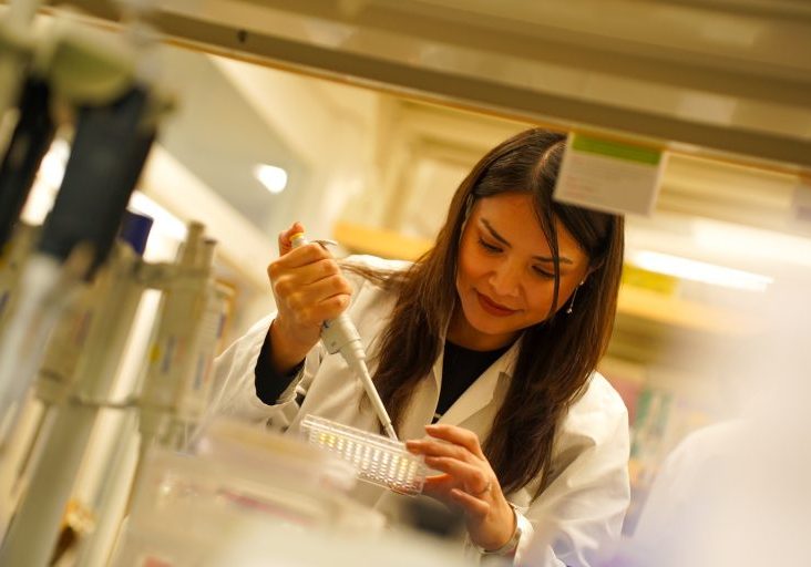 A researcher in a white lab coat uses a micropipette to transfer samples into a multiwell plate inside a laboratory.